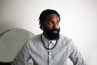 Dapper man in patterned dress shirt, bearded with dreadlocks, sitting thoughtfully.
